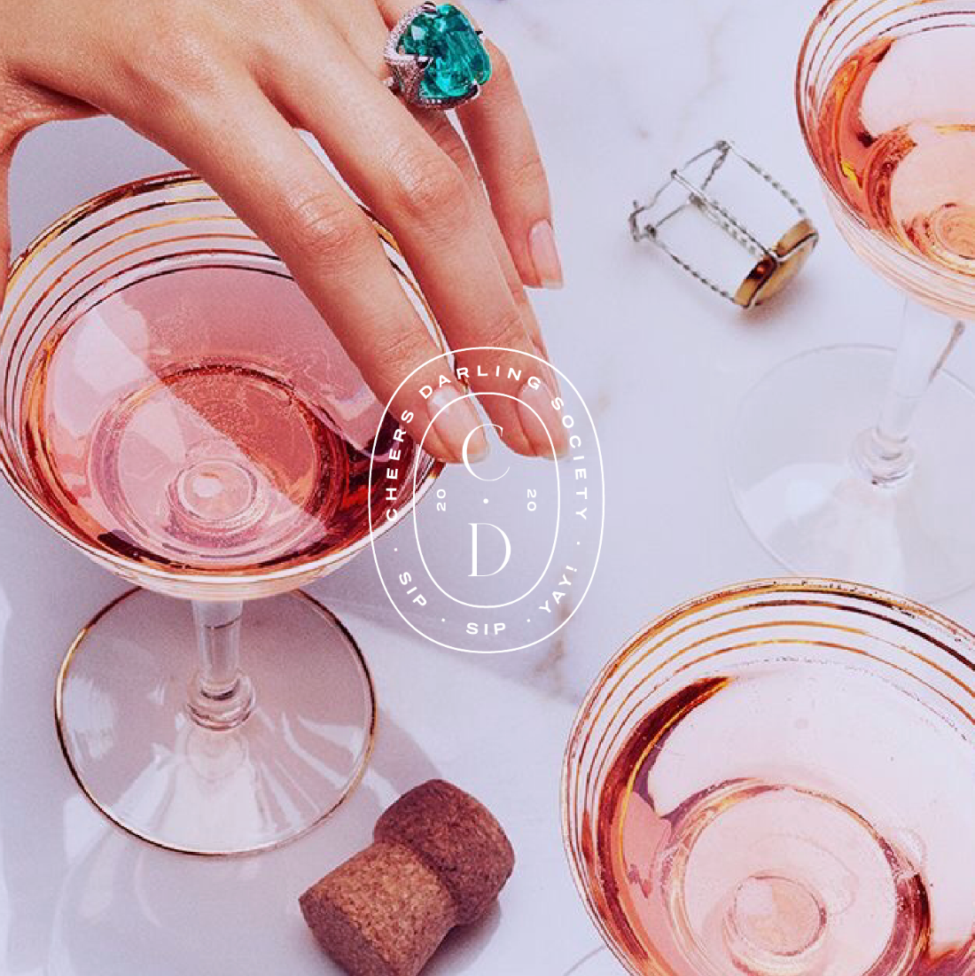 Hand with emerald ring holding a glass of pink champagne on a marble surface.